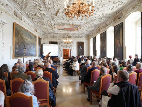 Blick auf einen Konzertsaal, in dem Personen auf Sesseln aus Holz und rotem Leder sitzen.