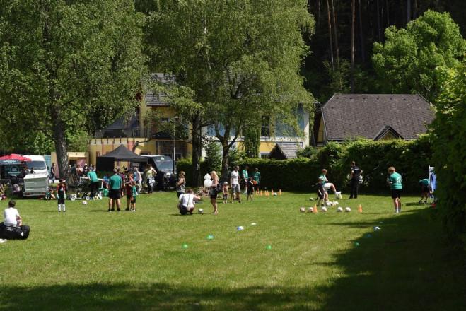 Erwachsene und Kinder spielen auf einer Wiese mit Bällen Fußball und weitere Personen stehen auf der Wiese.
