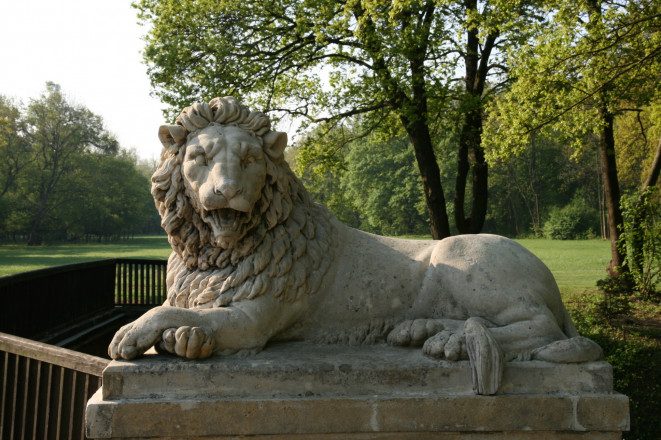 Blick auf eine Statue aus Stein von einem Löwen. Im Hintergrund sind Rasen und Bäume zu sehen.