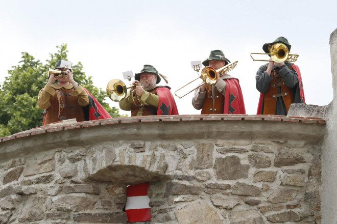 Vier Männer mit roten Umhängen und dunkelgrünen Hüten stehen hinter einer niedrigen Mauer und spielen Blasinstrumente.
