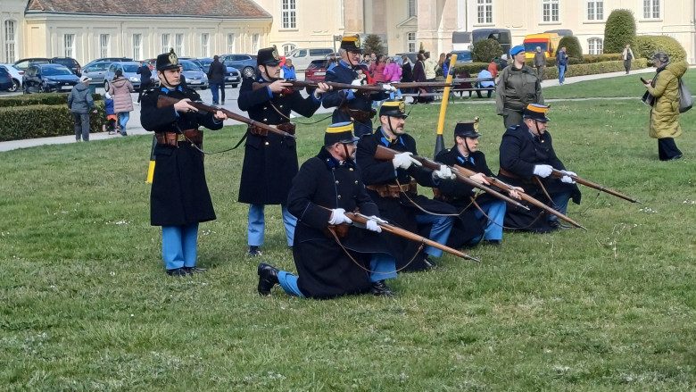 Männer im Kaiserlichen Gewand stehen mit Gewehren auf einer Wiese beisammen. Im Hintergrund ist das Schloss Laxenburg und ein Parkplatz zu sehen.