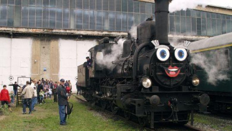 Blick auf eine Dampflock bei der vorne ein Gesicht aufgeklebt ist. Daneben stehen viele Personen.