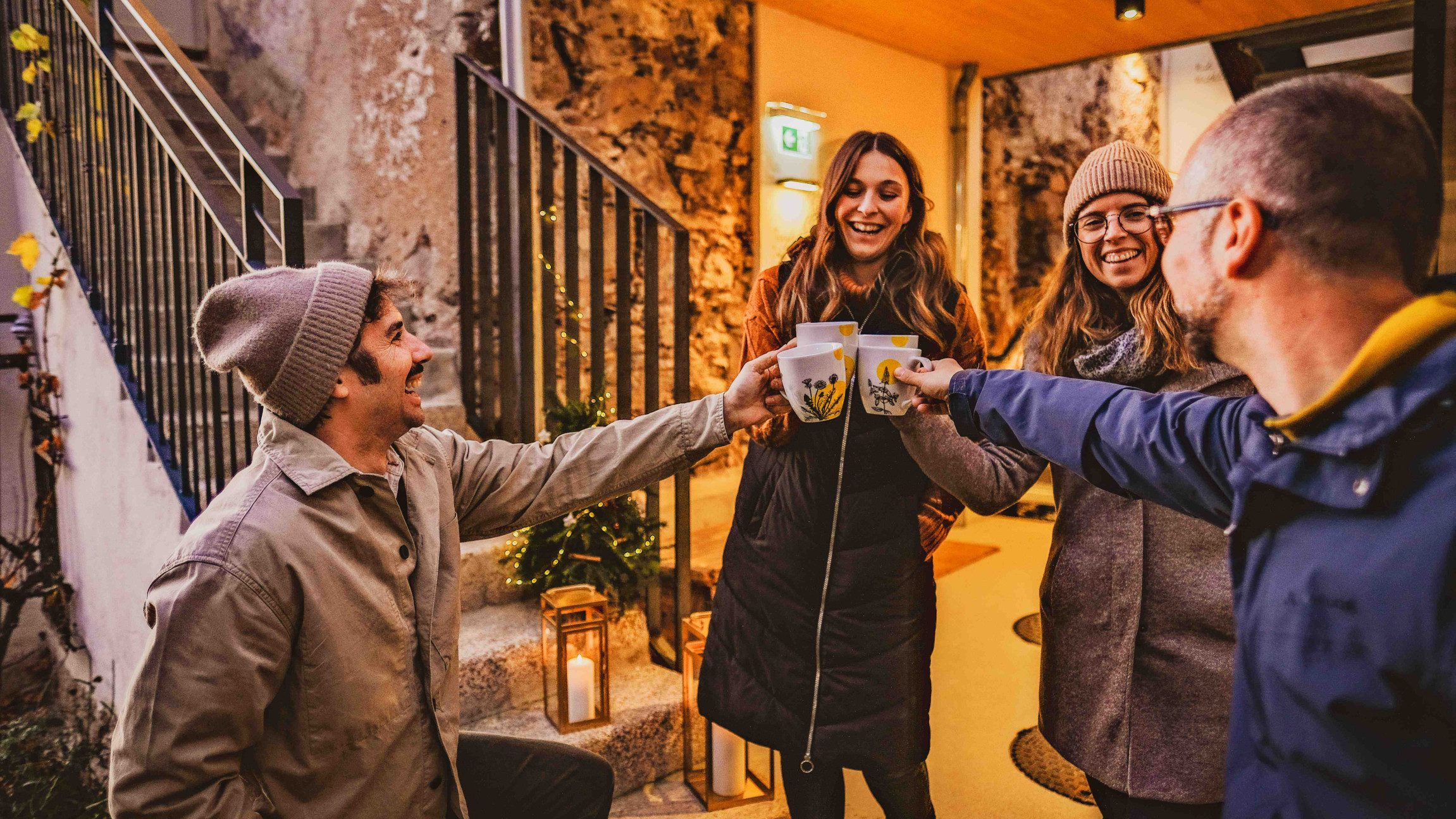 Vier winterlich gekleidete Personen stehen vor einer Treppe und einem Eingangsbereich und stoßen mit Tassen an.