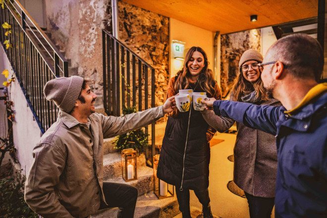 Vier winterlich gekleidete Personen stehen vor einer Treppe und einem Eingangsbereich und stoßen mit Tassen an.